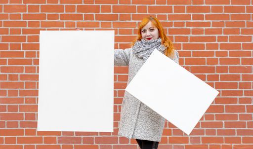 Young red-haired girl stands against the background of a red wall and holds two blank white canvases for painting in front of her