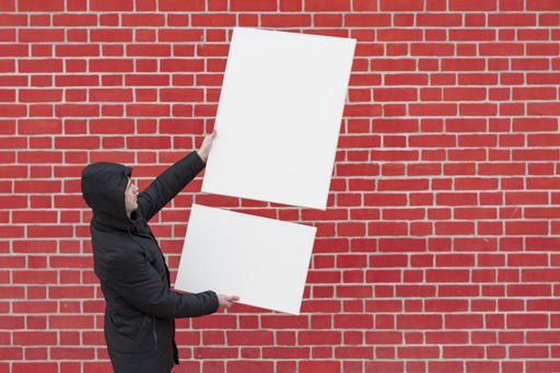 A young man stands against a background of a red brick wall and holds in front of him two white blank canvases for painting