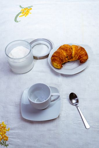 A top view closeup of a croissant on a white plate next to a container with powdered sugar and coffee