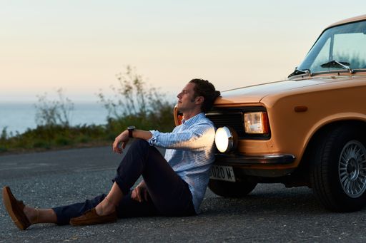 A beautiful shot of a man sitting in front of his vintage car looking at the lake near the street