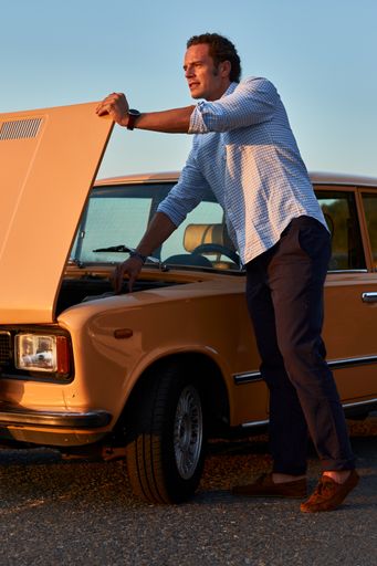 A vertical shot of a male model standing by his old vintage car with front side hood open