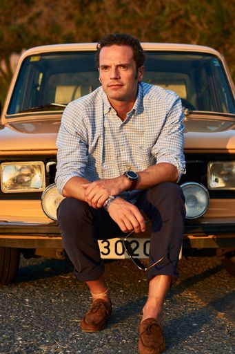 A vertical shot of a man sitting in front of his old vintage car. Serious look on a curly model face