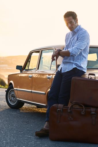 A vertical shot of a man standing near his vintage car with brown luggage holding his watch