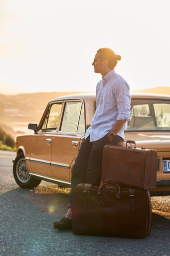 A vertical shot of man standing near his vintage car with brown luggage with tied hair and sunglasses