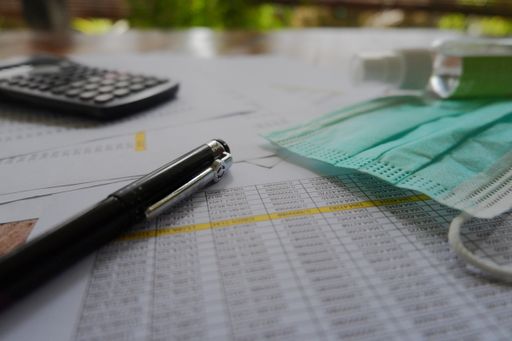 A closeup shot of a facemask, calculator, pen, hand sanitizer, and papers laying on the table
