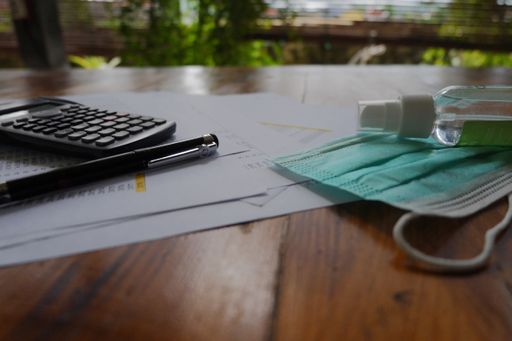 A closeup shot of a facemask, calculator, pen, hand sanitizer, and papers laying on the table