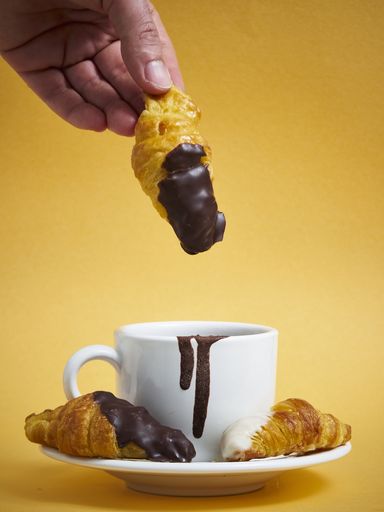 A closeup of a person dipping a croissant in the cup filled with chocolate on the yellow background