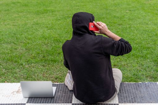 A young man wearing a hoodie sitting in a park with his laptop talking on the phone
