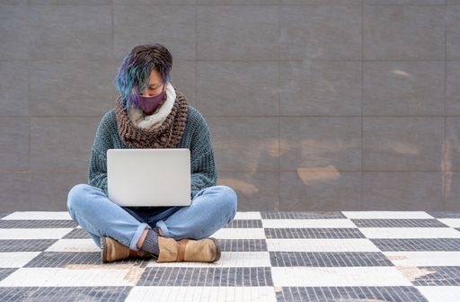A stylish young lady sitting on a checkered floor in a park using her laptop wearing a sanitary mask