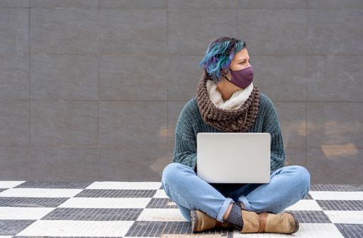 A stylish young lady sitting on a checkered floor in a park using her laptop wearing a sanitary mask