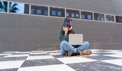 A stylish young lady sitting on a checkered floor in a park using her laptop talking on the phone wearing a sanitary mask