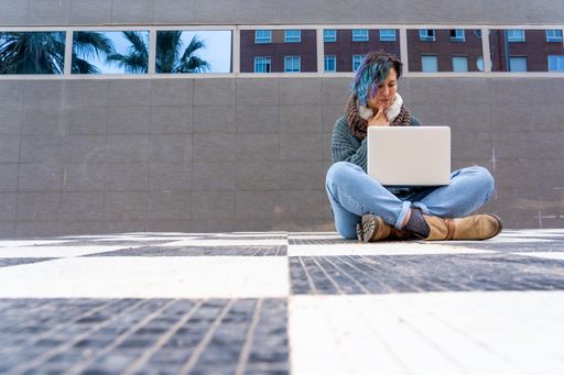 A stylish young lady with colorful hair sitting on a checkered floor in a park using her laptop