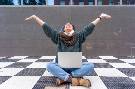 A cheerful young lady with colorful hair sitting on a checkered floor in a park using her laptop