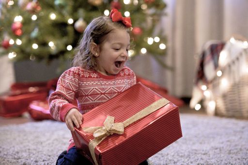 A little girl holding the Christmas gift in a red box with the Christmas tree on the background