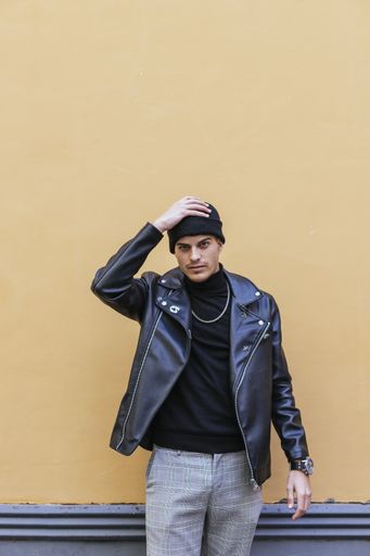 A vertical shot of a young man wearing leather coat with a turtleneck, necklace, trousers, and hat