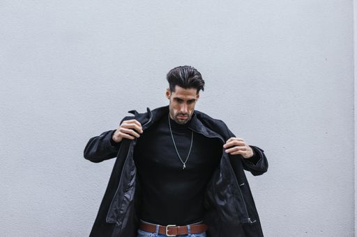 A closeup shot of a stylish man wearing a black coat posing in front of a gray wall