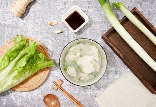 A top view closeup of a dumpling soup, next to lettuce leaves and soy sauce on a blue sur