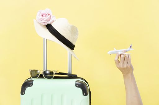 A closeup of a hand holding a toy airplane, sunglasses, hat, and luggage on a yellow background-concept travel