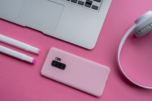 A high angle shot of a laptop, a cellphone, pens, and headphones on a pink surface