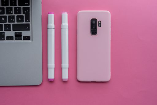 A high angle shot of a laptop, a pink cellphone, and pens on a pink surface