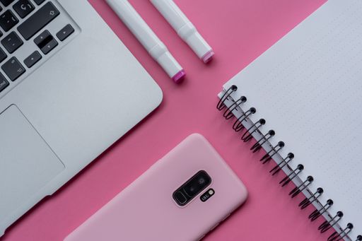 A high angle shot of a laptop, a cellphone, and writing tools on a pink surface