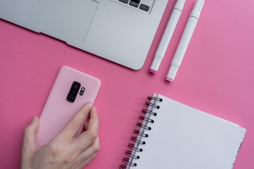 A top view of markers, notebook, laptop, and hand holding smartphone with pink case on pink background
