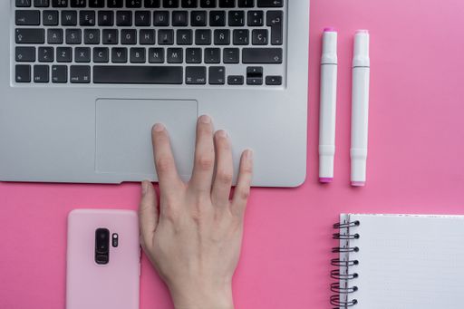 A high angle shot of a laptop, a cellphone, and writing tools on a pink surface