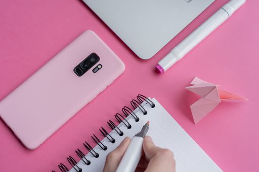 A high angle shot of a laptop, a cellphone, and writing tools on a pink surface