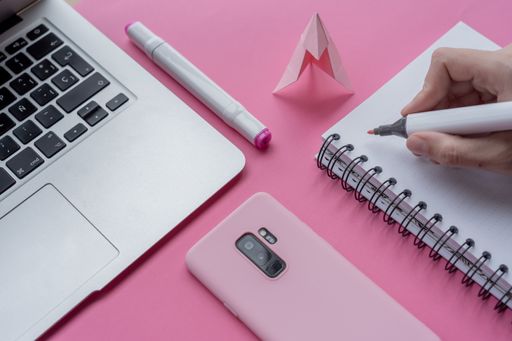 A hand holding marker with a notebook, laptop, phone, and swan origami on pink background