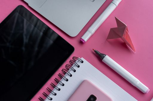 A high angle shot of a table, a cellphone, and writing tools on a pink surface
