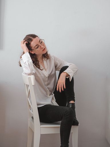 A vertical shot of a woman posing on the wooden chair on the background of a white wall
