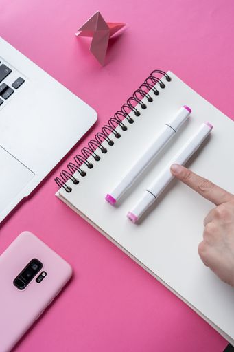 A vertical shot of a laptop, a cellphone, and writing tools on a pink surface