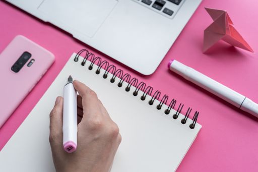 A high angle shot of a laptop, a cellphone, and writing tools on a pink surface