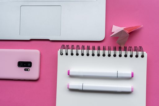 A  view of markers, notebook, laptop, phone, and swan origami on pink background