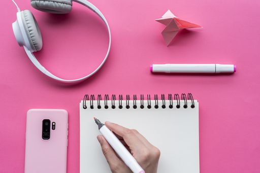 A top view of hand holding markers with a notebook, headphones, phone, and swan origami on pink background