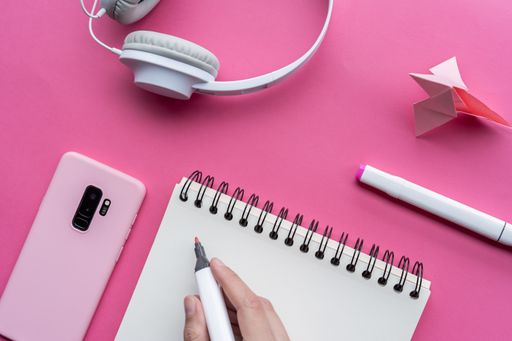 A high angle shot of a cellphone, headphones, and writing tools on a pink surface