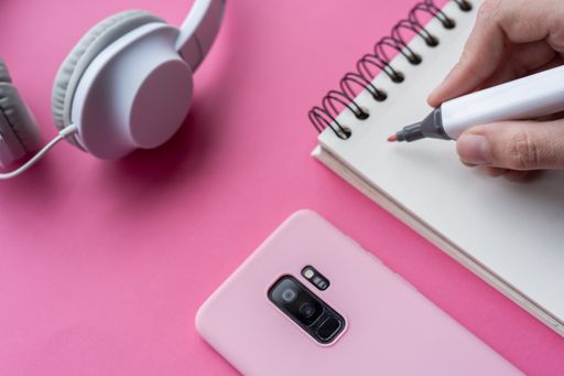 A top view of hand holding markers with a notebook, headphones, phone, and swan origami on pink background