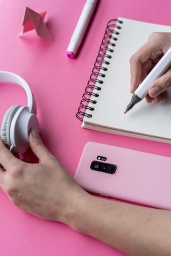 A of hand holding markers with a notebook, headphones, phone, and swan origami on pink background