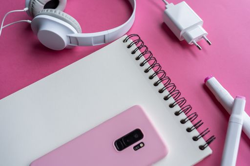 A high angle shot of a cellphone, headphones, and writing tools on a pink surface