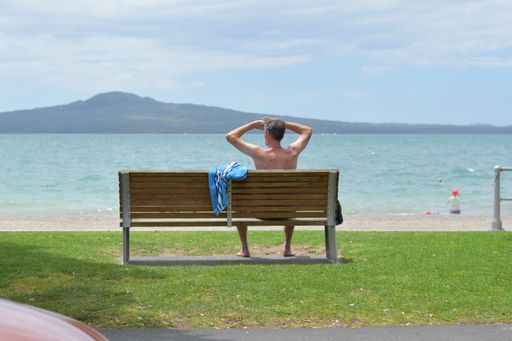 View of man sitting on bench, holding hands on his head and looking at Rangitoto dormant volcano island