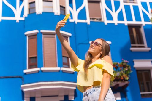 A young blonde caucasian female tourist taking selfies in a street with blue houses on a sunny day