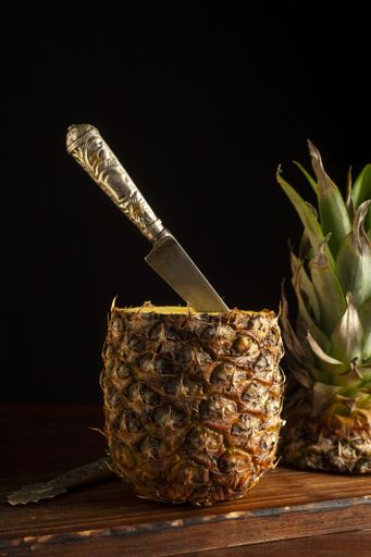 A vertical shot of a pineapple with a knife stuck in it and its cut crown on a wooden board