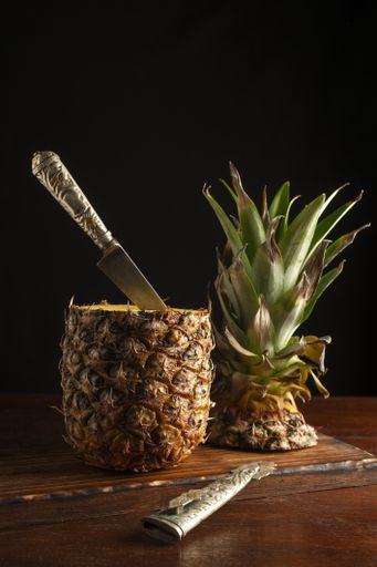 A vertical shot of a pineapple with a knife stuck in it and its cut crown on a wooden board