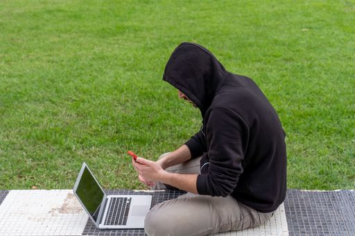 A closeup shot of a male in a hoodie sitting on a chess floor with a laptop and telephone in hands