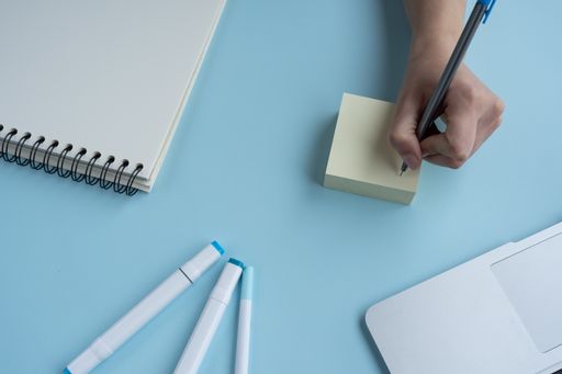 A person's hand writing on sticky notes on a blue desk with a laptop, notebook, and pens
