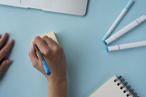 A person writing on sticky notes on a blue desk with a laptop, notebook, and pens