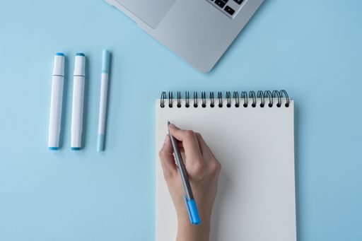 A person's hand writing in a notebook on a blue desk with a laptop and pen