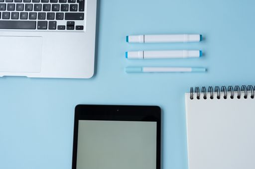 A high-angle shot of a blue desk with a laptop, pens, notebook and tablet