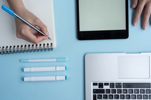 A high angle shot of a person taking notes using a tablet and a laptop on a blue desk
