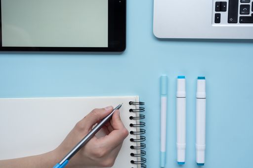 A high angle shot of a person taking notes using a tablet and a laptop on a blue desk